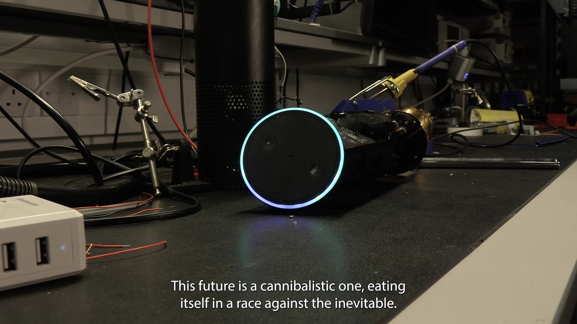 A shot from the film, showing amazon echo on a workbench with the subtitle 'this future is a cannablistic one, eating itself in a race against the inevitable'.